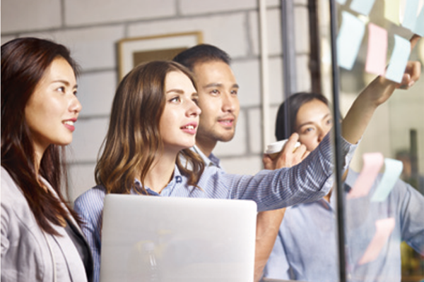 一群专业人士在工作空间中携手合作，积极参与讨论。一人指着贴满便利贴的玻璃板，另一人似乎在做笔记或使用笔记本电脑。气氛暗示了这是一个动态头脑风暴会议，从用户角度出发，增强设计感、促进可视化。